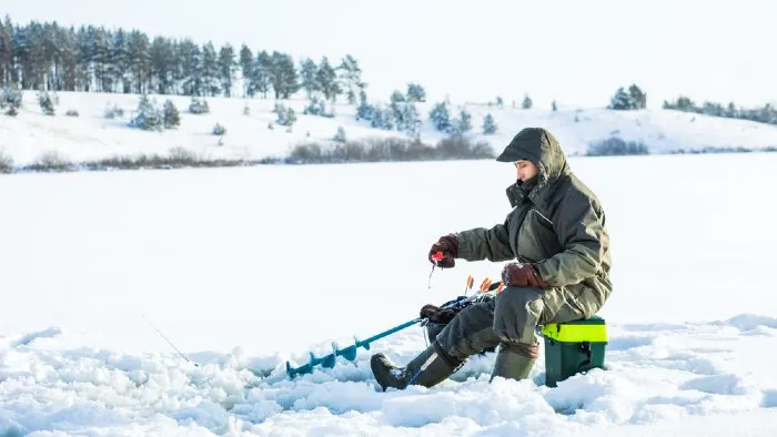 Man ice fishing with a tackle box, drill and pole.