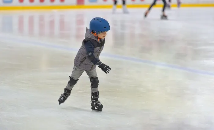 Adorable little boy in winter clothes with protections skating on ice rink

