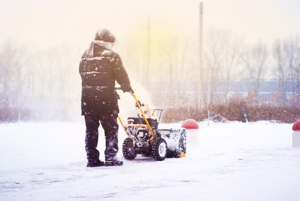 An adult pushes a snow blower through snow.