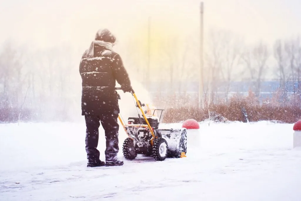 An adult pushes a snow blower through snow.
