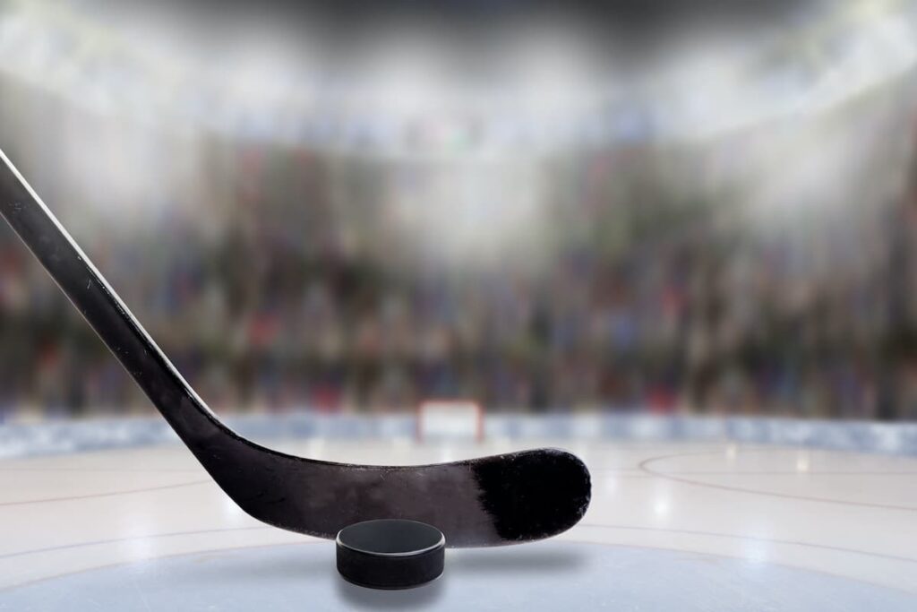 A hockey stick and puck on the ice with a blurred stadium crowd in the background.