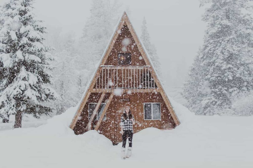 A young woman standing in front of an A-frame chalet cabin during a snowfall. Winter cabins in Minnesota.