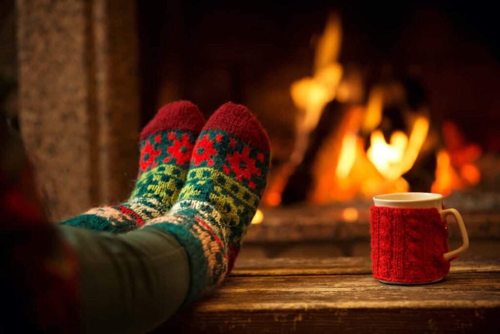 A single pair of feet in Christmas socks next to a mug covered in a knit warmer near a fireplace.