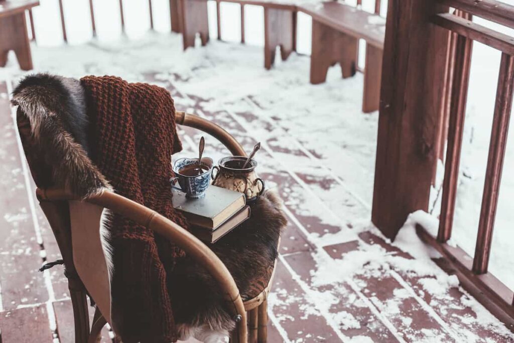 A chair with a blanket, books, and a cup of tea on a snowy cabin porch.